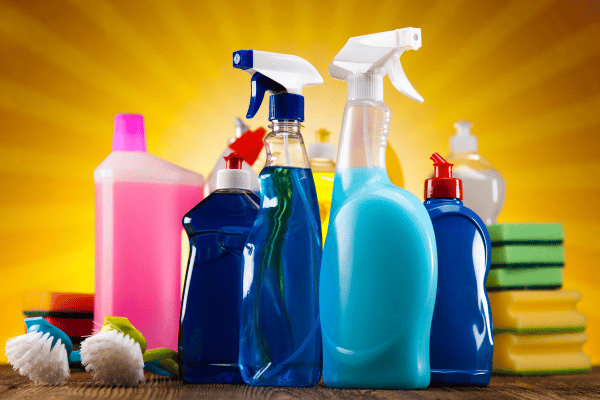 An assortment of colorful chemical cleaning products in spray bottles and containers, displayed alongside sponges and brushes against a bright background.