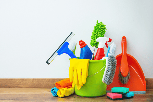 A bucket filled with various cleaning tools and supplies, including gloves, brushes, sponges, and spray bottles, placed on a wooden floor.