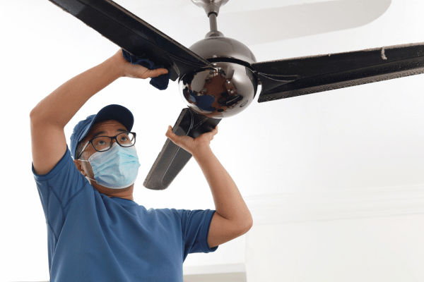 A person wearing a face mask and glasses cleaning a black ceiling fan with a cloth.
