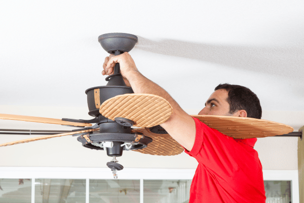 A man in a red shirt installing or adjusting a wooden-blade ceiling fan.