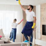 A team of professional cleaners wearing aprons and gloves is thoroughly cleaning a modern living room, with one person standing on a ladder to clean high surfaces.