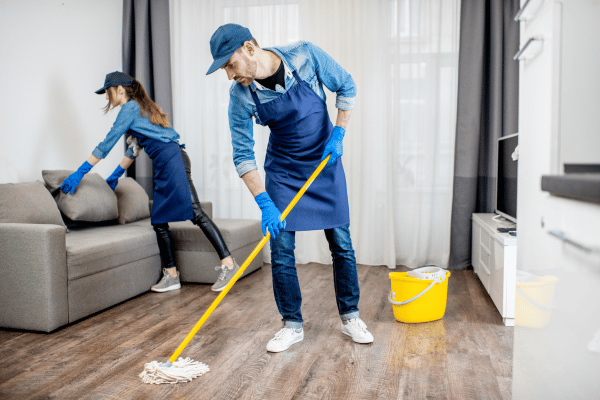 Two cleaners in uniforms are tidying up a living room, with one mopping the floor and the other arranging cushions on a couch.