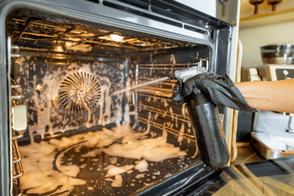 Close up of a gloved hand spraying cleaning solution inside a dirty oven.