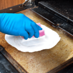 Close up of a hand with a blue glove cleaning an oven door with a pink sponge and white cleaning product.