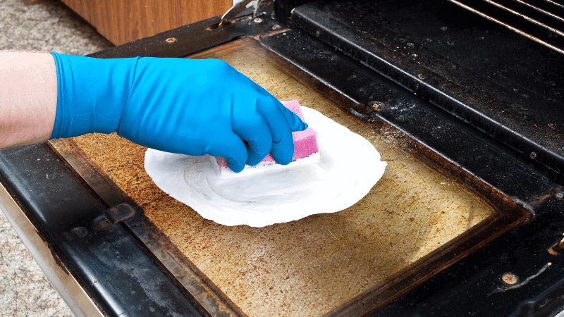 Close up of a hand with a blue glove cleaning an oven door with a pink sponge and white cleaning product.
