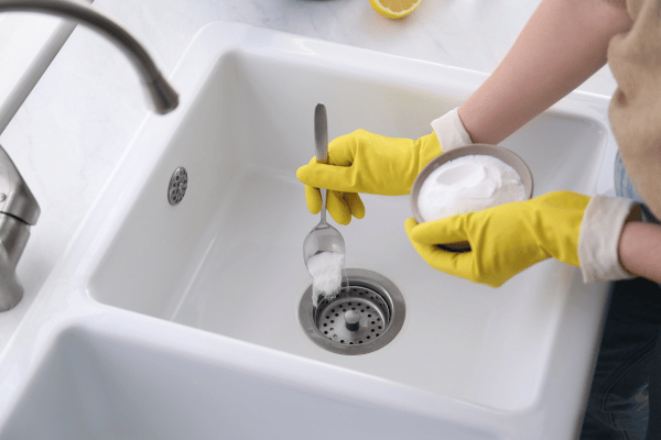 A person wearing yellow gloves pours baking soda into a kitchen sink drain for cleaning purposes.