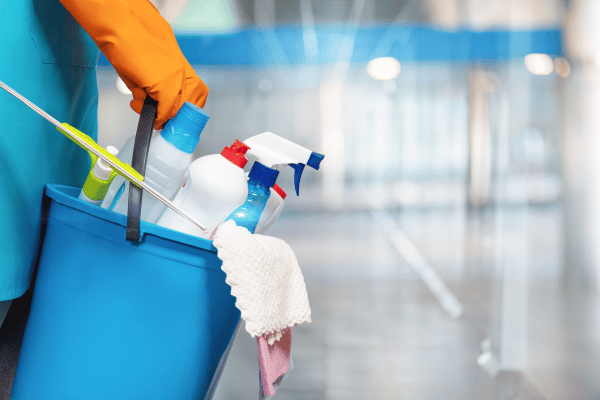 A person carrying a blue bucket filled with various cleaning supplies, including spray bottles, cloths, and brushes, in a brightly lit space.