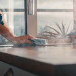 A person cleaning a kitchen countertop with a spray bottle and cloth under natural light.