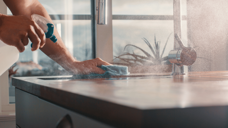 A person cleaning a kitchen countertop with a spray bottle and cloth under natural light.