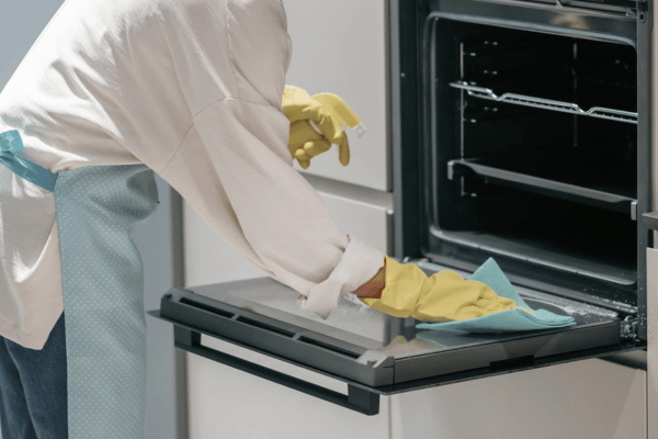 Person wearing yellow gloves and a blue apron cleaning the inside of an oven with a blue cloth.