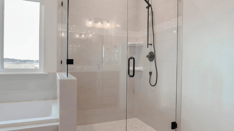 A modern glass-enclosed shower with a sleek black showerhead and handle, set against white tiled walls.