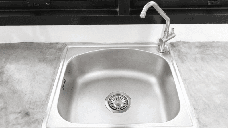 A stainless steel kitchen sink with a curved faucet, embedded in a polished concrete countertop.