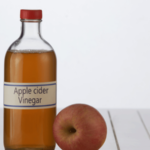 A bottle of apple cider vinegar with a red cap placed next to a fresh apple on a white wooden surface.