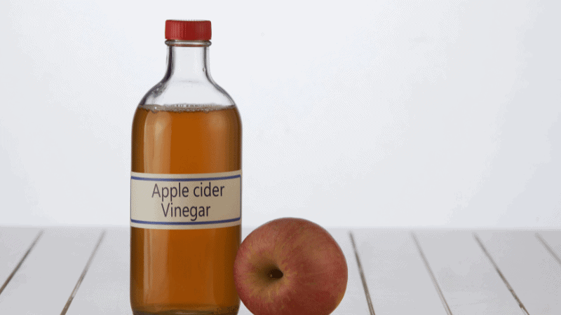 A bottle of apple cider vinegar with a red cap placed next to a fresh apple on a white wooden surface.