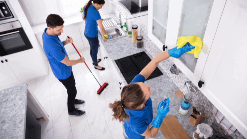 A team of cleaners working in a modern kitchen, wiping cabinets, mopping the floor, and organizing the space.