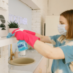 A person wearing pink gloves cleaning a white shelf in a well-lit kitchen with cleaning spray and a cloth.