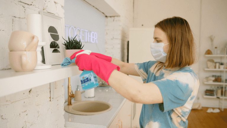 A person wearing pink gloves cleaning a white shelf in a well-lit kitchen with cleaning spray and a cloth.