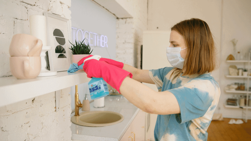 A person wearing pink gloves cleaning a white shelf in a well-lit kitchen with cleaning spray and a cloth.
