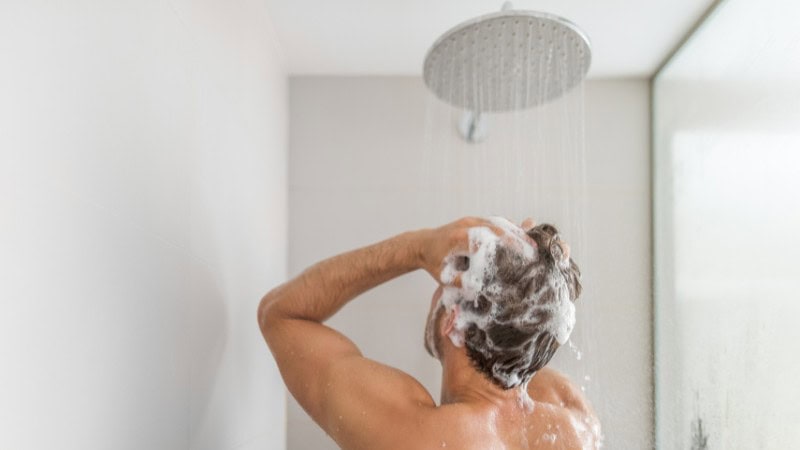 Person washing hair under shower with sudsy shampoo.