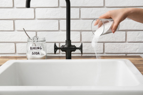 a hand is pouring baking soda all over the kitchen sink, a bottle of soda is near it.