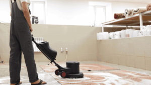 A worker uses a professional floor cleaning machine to wash large carpets inside a cleaning facility.