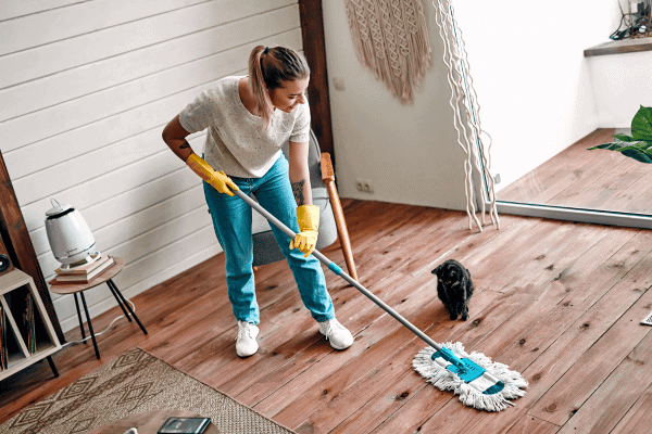 A woman wearing yellow cleaning gloves mops a wooden floor while a small black cat watches nearby.

