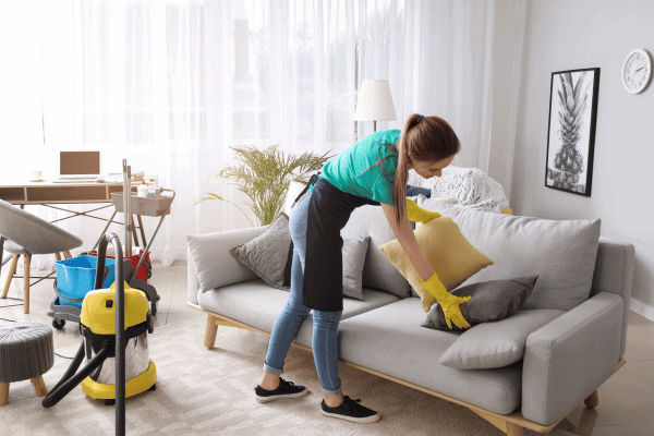 Woman wearing gloves arranging cushions while cleaning a bright living room.